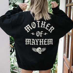 May include: Black sweatshirt featuring the white text "MOTHER OF MAYHEM" in a gothic font, a lightning bolt, and a skull with wings. The sweatshirt is worn by a person standing in a doorway.