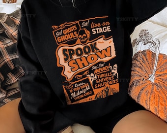 May include: Black Halloween-themed sweatshirt with orange and white text that reads "SPOOK SHOW." The design includes a skull, ghosts, and the phrases "See! Ghosts, Ghouls," and "Live on Stage." A pumpkin-shaped pillow is visible.