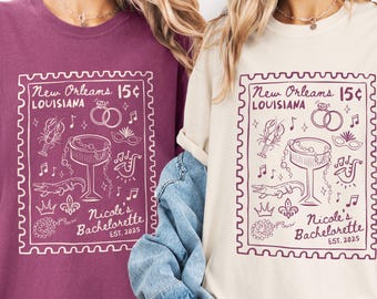 May include: Two t-shirts, one plum and one cream, each featuring a stamp-like design with the text "New Orleans 15¢ LOUISIANA" and "Nicole's Bachelorette EST. 2025". The design includes illustrations of a crawfish, rings, and musical notes.