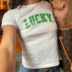 May include: White t-shirt with the word "LUCKY" printed in green block letters. The shirt is paired with blue jeans. A brown handbag with a monogram pattern is visible.
