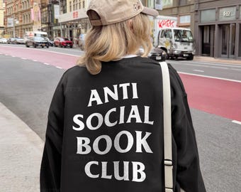 Puede incluir: Sudadera negra con el texto blanco "ANTI SOCIAL BOOK CLUB". La persona lleva una gorra de béisbol beige y un bolso blanco. El fondo muestra una calle de la ciudad con coches y edificios.