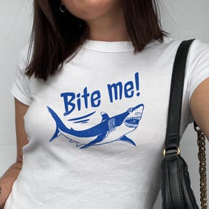 Puede incluir: Camiseta blanca con un gr&aacute;fico de tibur&oacute;n azul y el texto "Bite me!". La ilustraci&oacute;n del tibur&oacute;n presenta dientes afilados y se encuentra sobre un fondo blanco. La camiseta es de cuello redondo.