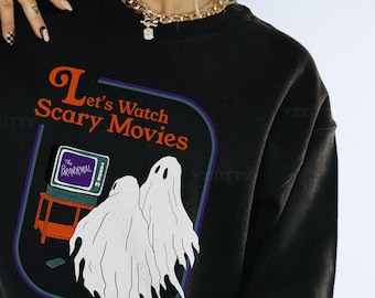 May include: Black sweatshirt with a graphic that reads "Let's Watch Scary Movies." The design features two white ghost figures, a television, and the text "The Paranormal." The design is framed in a purple and green border.