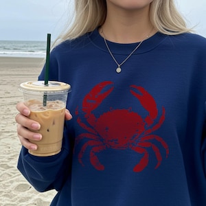May include: Navy blue sweatshirt featuring a large red crab graphic. The sweatshirt is a crew neck style and appears to be made of a soft material. The image is taken on a beach, with a person holding a beverage.