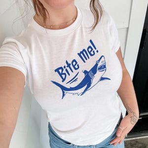 Puede incluir: Camiseta blanca con un dise&ntilde;o azul de un tibur&oacute;n y la frase "Bite me!" en una fuente llamativa. El tibur&oacute;n se representa con dientes afilados y una expresi&oacute;n amenazante. Una prenda casual para el uso diario.