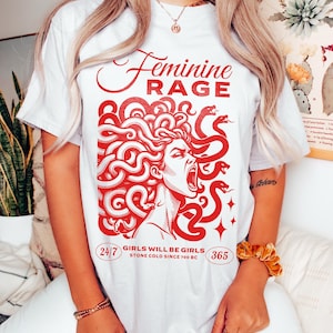May include: White t-shirt with a red graphic of a woman with snakes for hair. The text "Feminine Rage" is above the graphic and "Girls will be girls, stone cold since 700 BC" is below the graphic. The numbers 24/7 and 365 are on either side of the text.