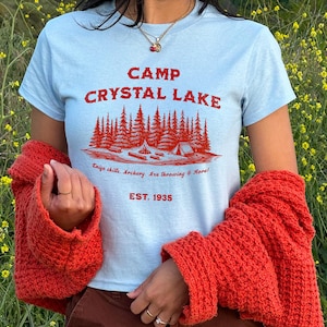 May include: A light blue t-shirt with a red graphic print that says "Camp Crystal Lake" with a forest scene and the text "Enjoy chills, archery, axe throwing & more! Est. 1935".