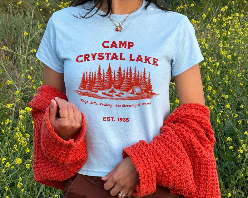 May include: A light blue t-shirt with a red graphic print that says "Camp Crystal Lake" with a forest scene and the text "Enjoy chills, archery, axe throwing & more! Est. 1935".