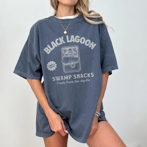 May include: A faded blue t-shirt with the text "BLACK LAGOON" above a graphic of a swamp creature in a can. Below the graphic, the text reads "SWAMP SNACKS Fresh From the Depths". The shirt has short sleeves and a relaxed fit.
