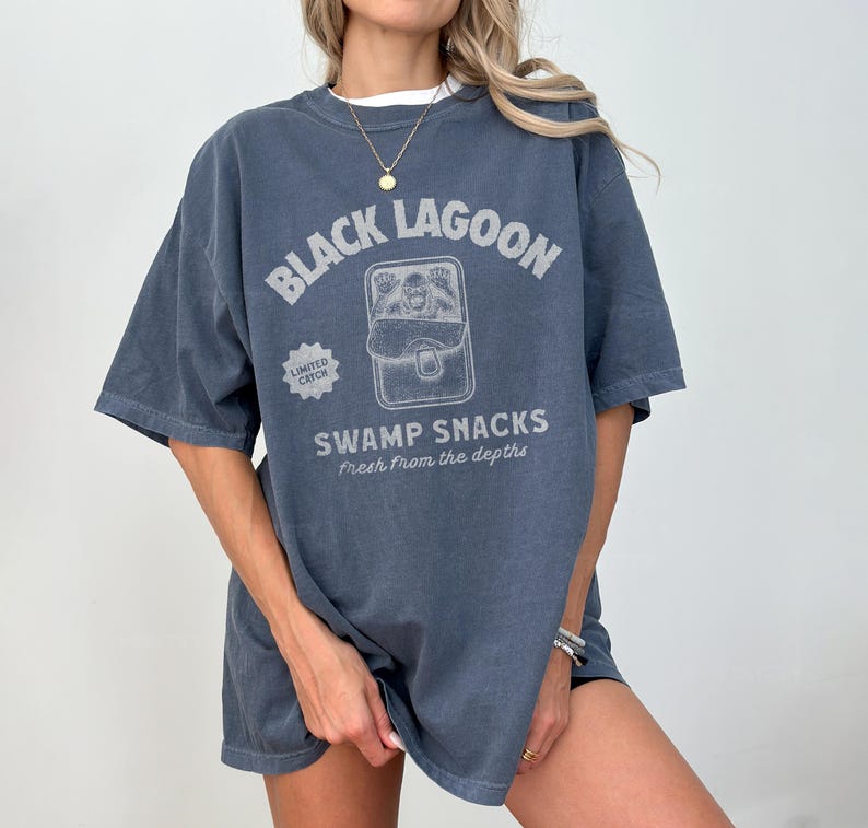 May include: A faded blue t-shirt with the text "BLACK LAGOON" above a graphic of a swamp creature in a can. Below the graphic, the text reads "SWAMP SNACKS Fresh From the Depths". The shirt has short sleeves and a relaxed fit.