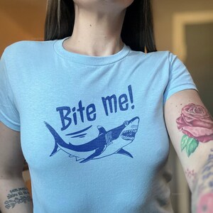 Puede incluir: Camiseta azul claro con un dise&ntilde;o de tibur&oacute;n azul marino y el texto "Bite me!". El tibur&oacute;n tiene dientes afilados y una forma curvada. La camiseta es de cuello redondo.