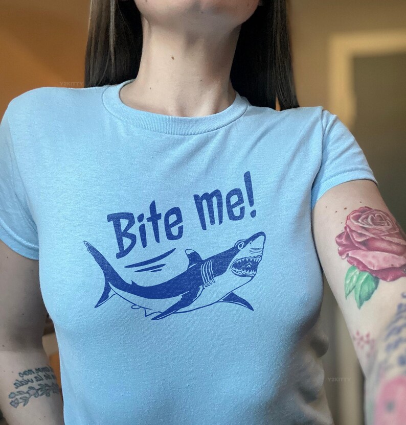 Puede incluir: Camiseta azul claro con un dise&ntilde;o de tibur&oacute;n azul marino y el texto "Bite me!". El tibur&oacute;n tiene dientes afilados y una forma curvada. La camiseta es de cuello redondo.