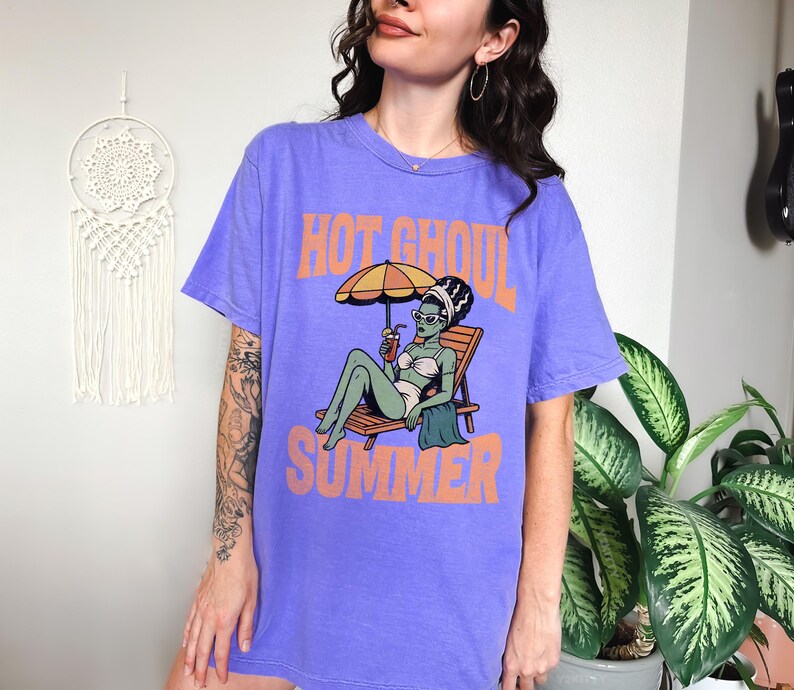 May include: A purple short-sleeve t-shirt featuring a graphic of a green-skinned figure in a white bikini lounging on a wooden chair under an orange umbrella, holding a drink. The text "HOT GHOUL SUMMER" is printed in orange above and below the graphic.