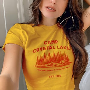 Puede incluir: Camiseta amarilla de manga corta con un gráfico rojo que dice "Camp Crystal Lake" con un dibujo de una escena de bosque y el texto "Knife skills, Archery, Axe throwing & More! EST. 1935"