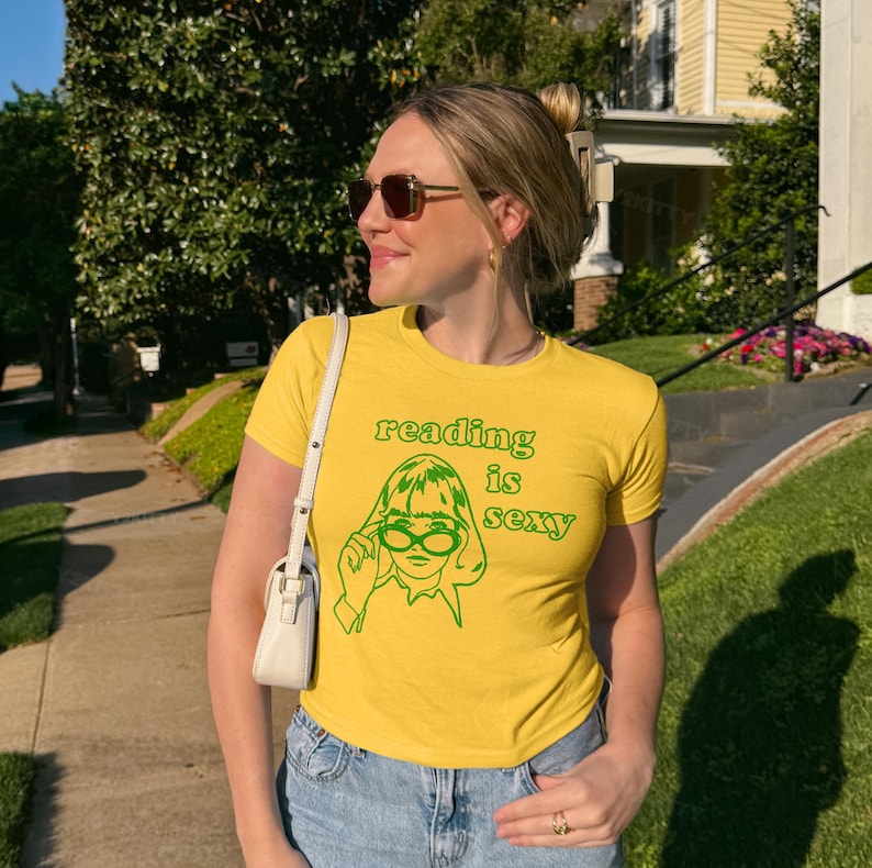 May include: Yellow t-shirt with the text "reading is sexy" and a green graphic of a woman wearing glasses. The shirt is paired with blue jeans and a white purse. The person is wearing sunglasses and has their hair up.