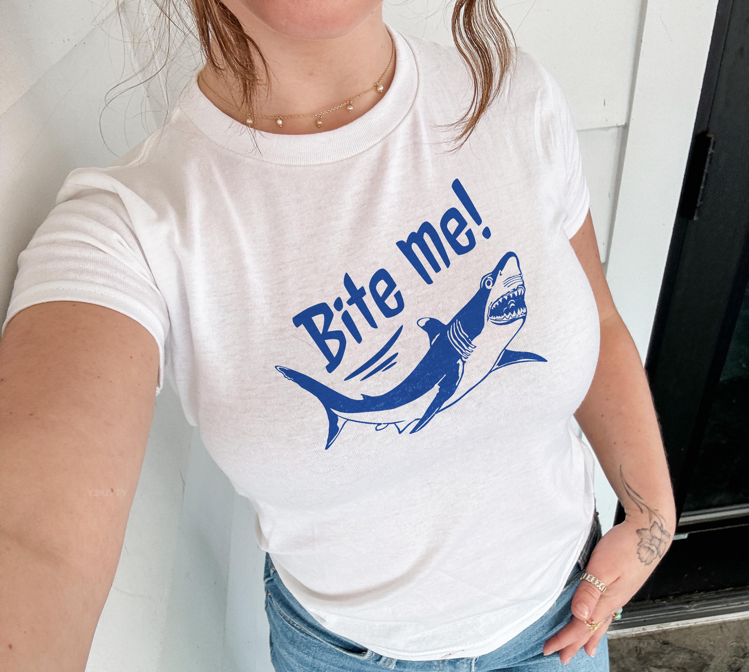Puede incluir: Camiseta blanca con un dise&ntilde;o azul de un tibur&oacute;n y la frase "Bite me!" en una fuente llamativa. El tibur&oacute;n se representa con dientes afilados y una expresi&oacute;n amenazante. Una prenda casual para el uso diario.