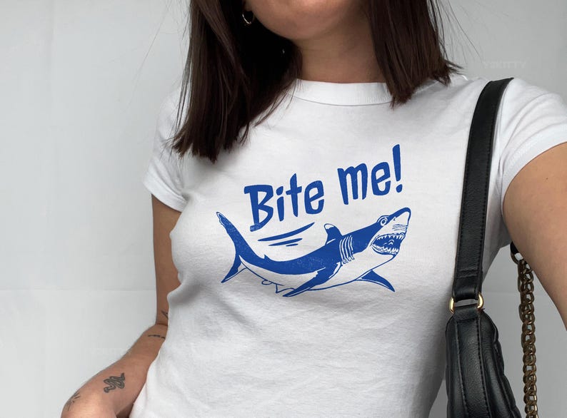 Puede incluir: Camiseta blanca con un gr&aacute;fico de tibur&oacute;n azul y el texto "Bite me!". La ilustraci&oacute;n del tibur&oacute;n presenta dientes afilados y se encuentra sobre un fondo blanco. La camiseta es de cuello redondo.