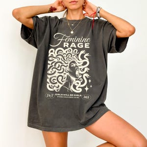May include: A charcoal grey oversized t-shirt with a graphic print of a woman with snakes for hair. The text "Feminine Rage" is printed above the graphic. The text "Girls will be girls. Stone cold since 700 BC" is printed below the graphic.