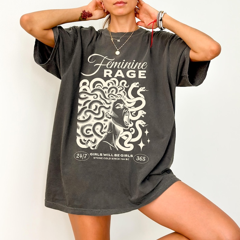 May include: A charcoal grey oversized t-shirt with a graphic print of a woman with snakes for hair. The text "Feminine Rage" is printed above the graphic. The text "Girls will be girls. Stone cold since 700 BC" is printed below the graphic.