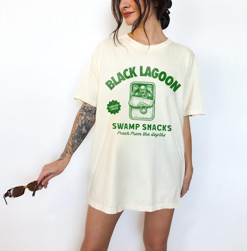 May include: Cream-colored t-shirt with a green graphic that reads "Black Lagoon" above an illustration of a creature in a can labeled "Swamp Snacks." The text "Fresh from the depths" is below the can. A "Limited Catch" badge is on the left.