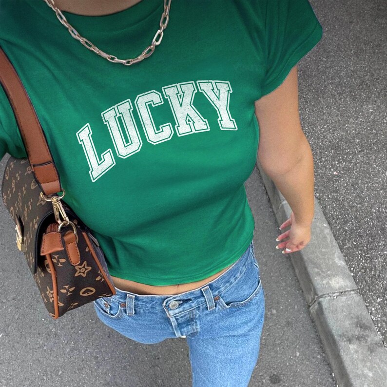 May include: A green t-shirt with the word "LUCKY" in white block letters. The shirt is paired with light blue jeans and a brown purse with a gold chain strap. A silver chain necklace is also visible.
