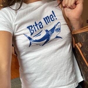 Puede incluir: Camiseta blanca con el texto "Bite me!" en azul, con un gr&aacute;fico de tibur&oacute;n azul. La camiseta es de cuello redondo y se combina con vaqueros azules.