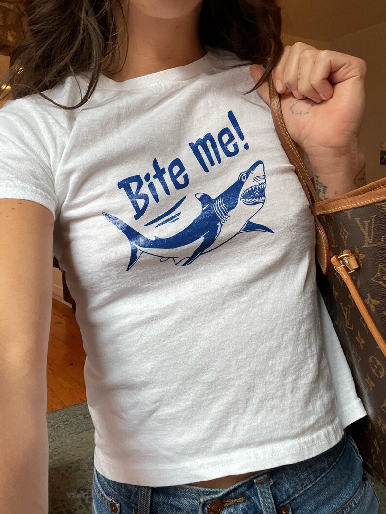 Puede incluir: Camiseta blanca con el texto "Bite me!" en azul, con un gr&aacute;fico de tibur&oacute;n azul. La camiseta es de cuello redondo y se combina con vaqueros azules.