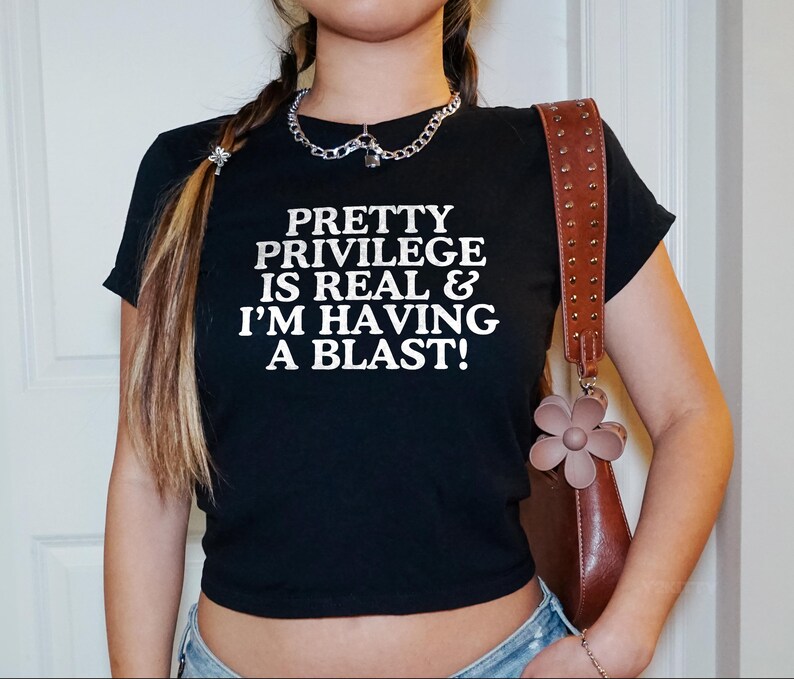 May include: A black cropped t-shirt with the text "Pretty Privilege is Real & I'm Having A Blast!" in white lettering. The shirt is being worn by a person with long brown hair and a silver chain necklace.