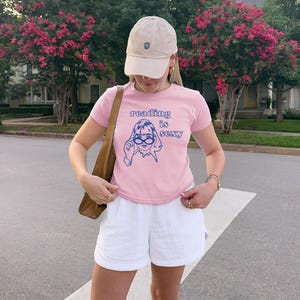 May include: A light pink t-shirt with the text "reading is sexy" and a blue graphic of a woman wearing glasses. The shirt is paired with white shorts and a beige baseball cap.