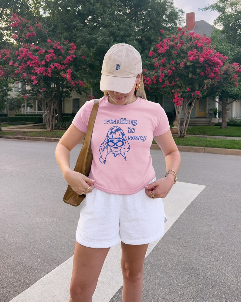 May include: A light pink t-shirt with the text "reading is sexy" and a blue graphic of a woman wearing glasses. The shirt is paired with white shorts and a beige baseball cap.