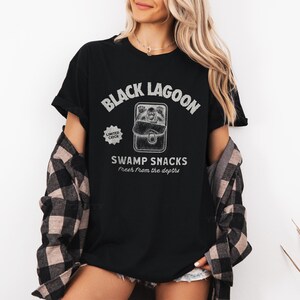 May include: Black t-shirt with the text "Black Lagoon" above a graphic of a creature in a lunchbox, with the words "Swamp Snacks" and "Fresh from the depths" below. The shirt also has a "Limited Catch" graphic.