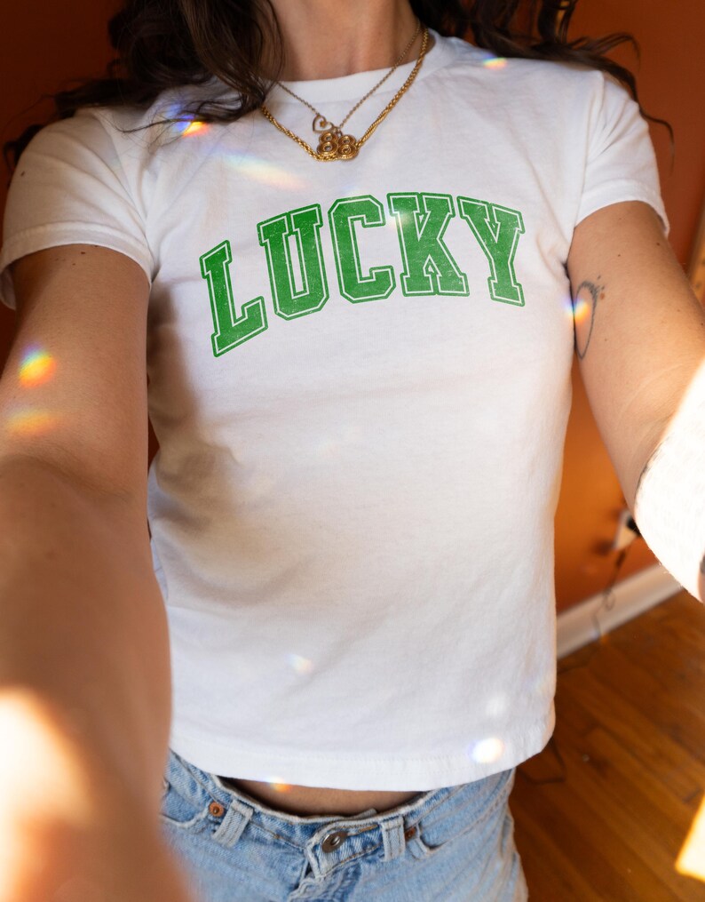 May include: White t-shirt with the word "LUCKY" printed in green block letters. The shirt has a crew neck and short sleeves. The model is wearing a gold necklace and blue jeans.