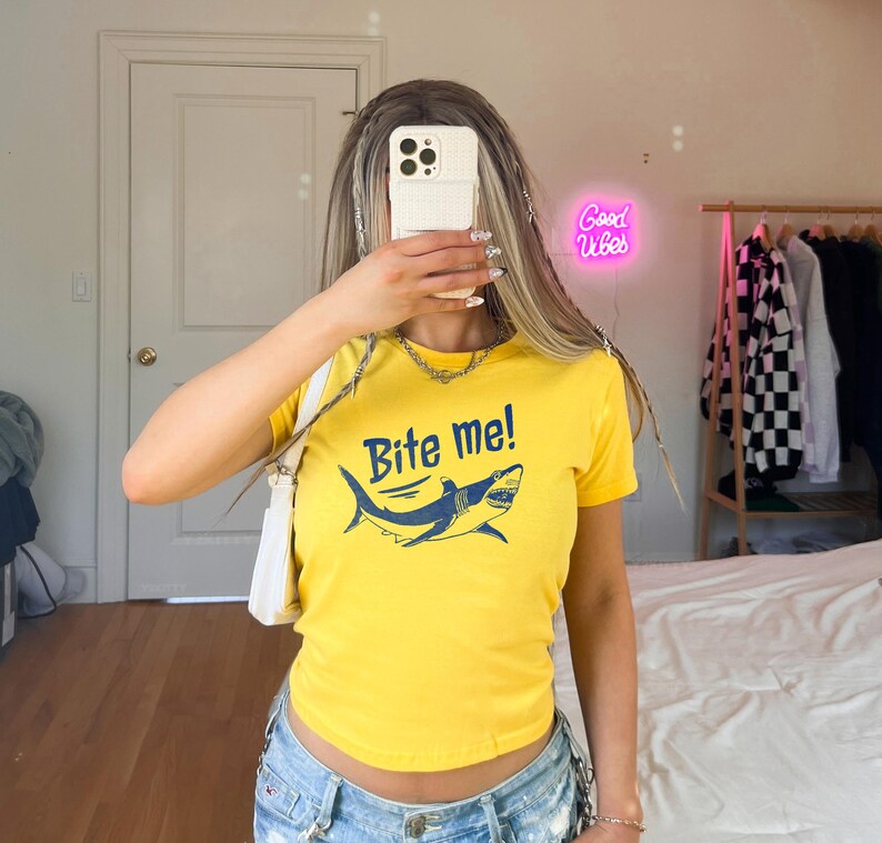 Puede incluir: Camiseta amarilla con el texto "Bite me!" y un gr&aacute;fico de tibur&oacute;n azul. La camiseta de manga corta tiene cuello redondo. La camiseta est&aacute; hecha de un material suave.