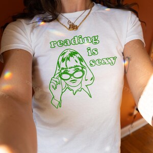 May include: White t-shirt with a green graphic that says "reading is sexy." The graphic features a line drawing of a person wearing glasses. The shirt is a crew neck and appears to be made of cotton.