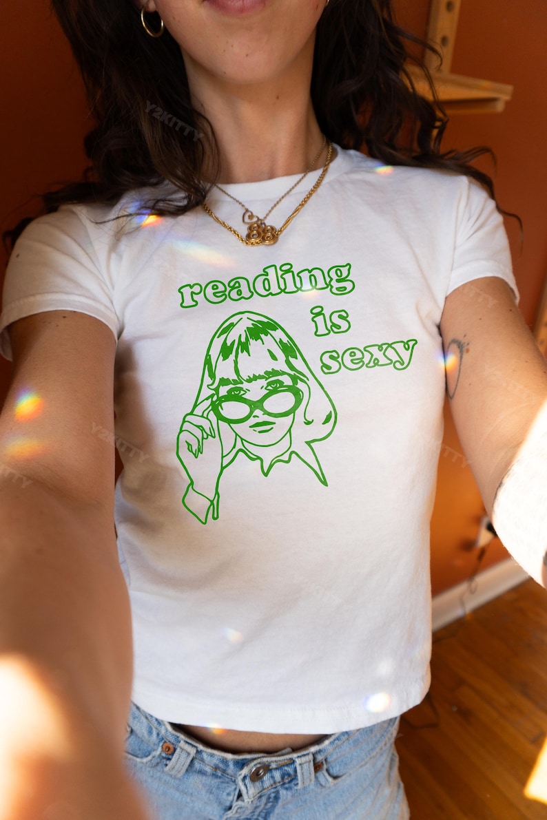 May include: White t-shirt with a green graphic that says "reading is sexy." The graphic features a line drawing of a person wearing glasses. The shirt is a crew neck and appears to be made of cotton.