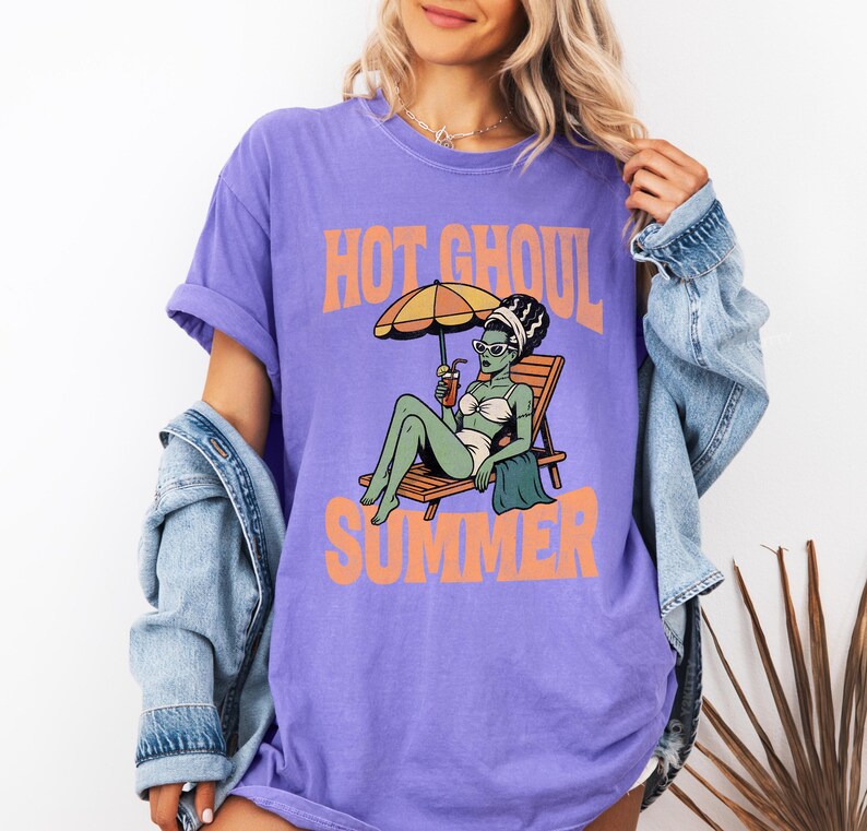 May include: A purple short-sleeve t-shirt featuring a graphic of a green-skinned ghoul in a white bikini lounging on a beach chair under an umbrella, holding a drink. The text HOT GHOUL SUMMER is printed in orange above and below the illustration.
