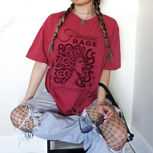 May include: A red t-shirt with a graphic of a woman with snakes in her hair. The text "Feminine Rage" is printed on the shirt. The text "Girls will be girls, stone cold since forever" is printed below the graphic.
