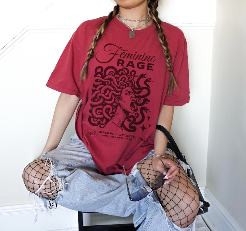 May include: A red t-shirt with a graphic of a woman with snakes in her hair. The text "Feminine Rage" is printed on the shirt. The text "Girls will be girls, stone cold since forever" is printed below the graphic.