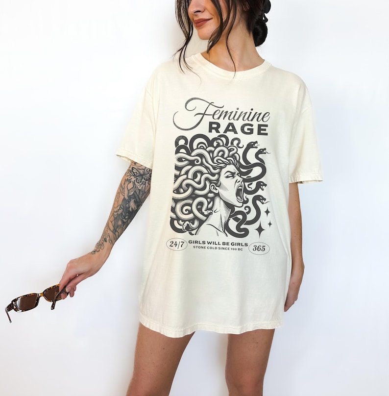 May include: A cream-colored t-shirt with a black and white graphic of a woman with snakes for hair. The text "Feminine Rage" is above the graphic and "Girls will be girls. Stone cold since 700 BC" is below the graphic.