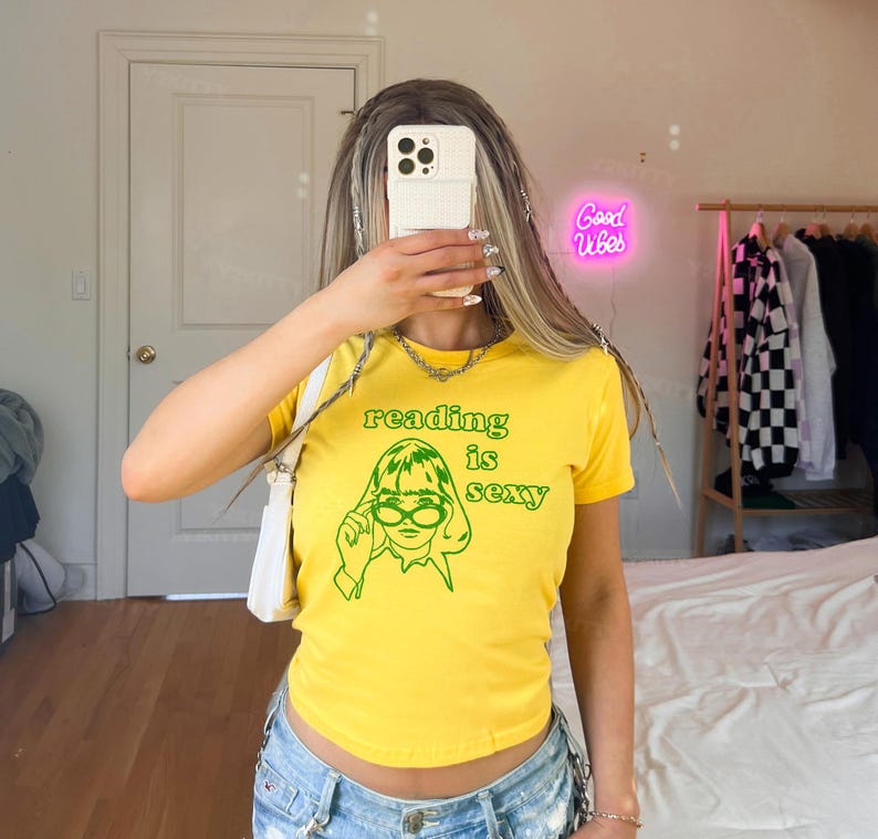 May include: Yellow t-shirt with the text "reading is sexy" in green, along with a green graphic of a woman wearing glasses. The shirt is short-sleeved and has a scoop neck.