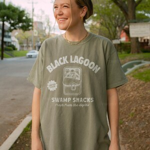May include: Olive green t-shirt with the text "BLACK LAGOON" above a graphic of a creature and the words "SWAMP SNACKS fresh from the depths." A "LIMITED CATCH" logo is on the left. The shirt is worn by a person.