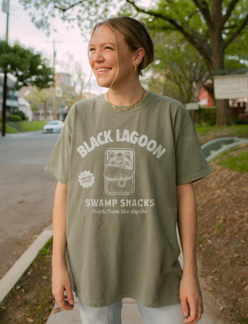 May include: Olive green t-shirt with the text "BLACK LAGOON" above a graphic of a creature and the words "SWAMP SNACKS fresh from the depths." A "LIMITED CATCH" logo is on the left. The shirt is worn by a person.