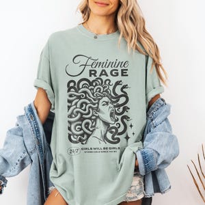 May include: A light green t-shirt with a graphic print of a woman with snakes for hair. The text "Feminine Rage" is printed above the graphic. The text "Girls will be girls. Stone cold since 70 BC" is printed below the graphic.