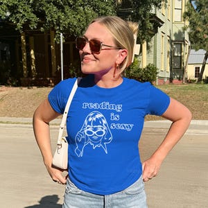 May include: A royal blue t-shirt with the text "reading is sexy" and a white graphic of a woman wearing glasses. The shirt is paired with denim shorts and a white purse.