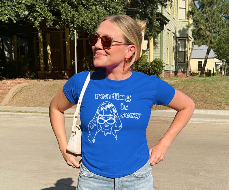 May include: A royal blue t-shirt with the text "reading is sexy" and a white graphic of a woman wearing glasses. The shirt is paired with denim shorts and a white purse.