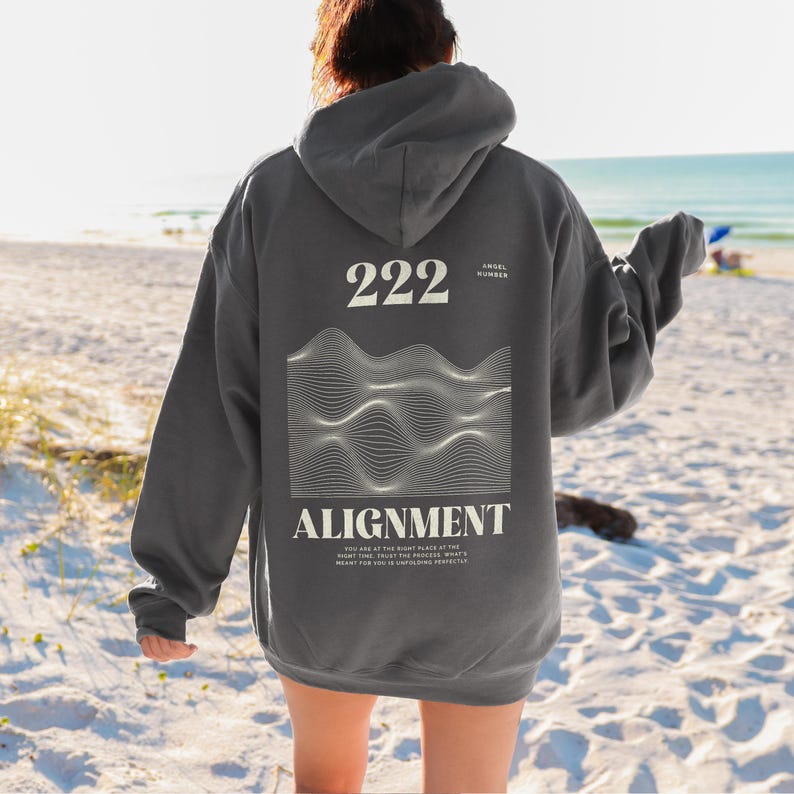 May include: A charcoal grey hooded sweatshirt with a white graphic design on the back. The graphic features a wavy line pattern and the text "222 Angel Number Alignment You are at the right place at the right time. Trust the process. What's meant for you is coming perfectly."