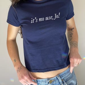 May include: Navy blue crop top with the text "it's no use, Jo!" in white script. The shirt has short sleeves and a crew neck. The model is wearing blue jeans.