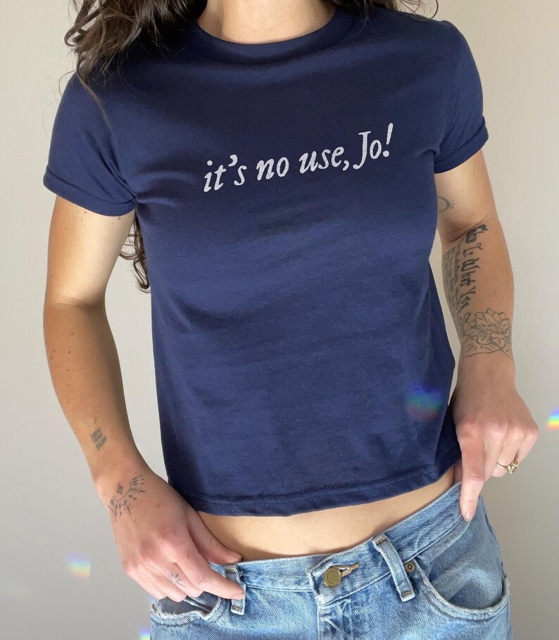 May include: Navy blue crop top with the text "it's no use, Jo!" in white script. The shirt has short sleeves and a crew neck. The model is wearing blue jeans.