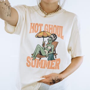 May include: An off-white short-sleeve t-shirt featuring a graphic of a green-skinned character in a white bikini and sunglasses lounging on a beach chair under an umbrella with a drink. The text reads HOT GHOUL SUMMER in orange letters.