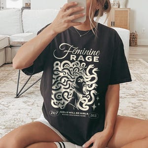May include: Black t-shirt with a white graphic of a woman with snakes for hair. The text "Feminine Rage" is above the graphic and "Girls will be girls. Stone cold since 700 BC" is below the graphic.